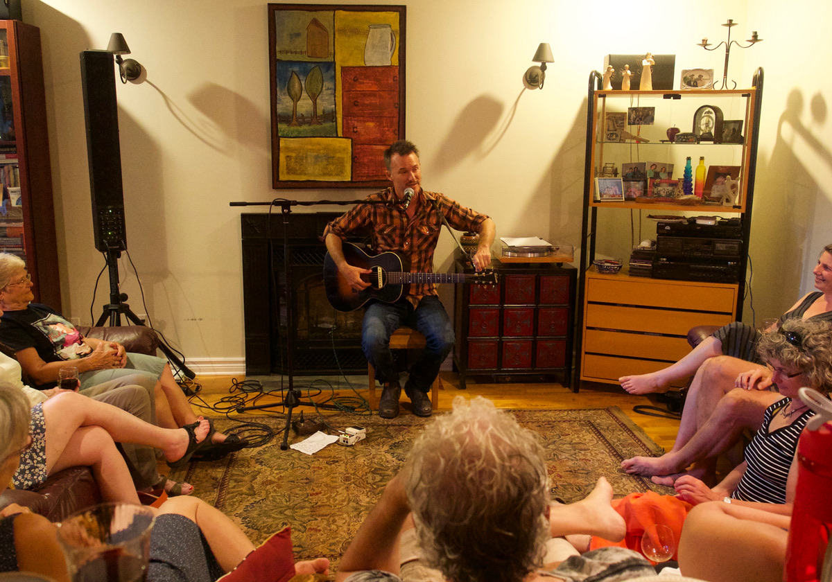 A musician playing guitar in a living room full of seated listeners